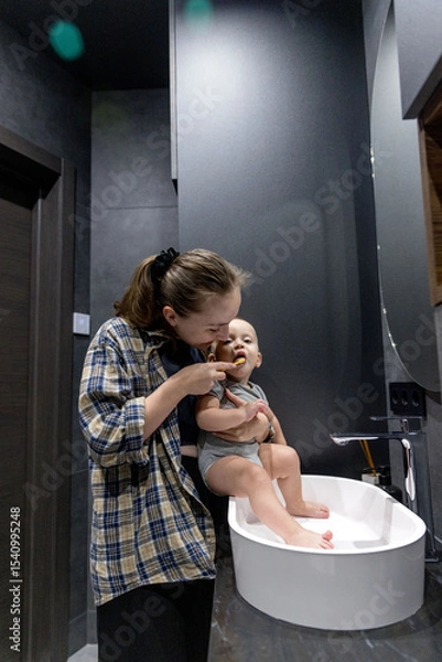 Fototapeta A woman with her baby brushes her teeth while holding a baby in hands in the bathroom