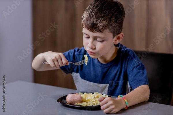 Fototapeta Little boy is happily eating pasta farfalle with sausage in the kitchen