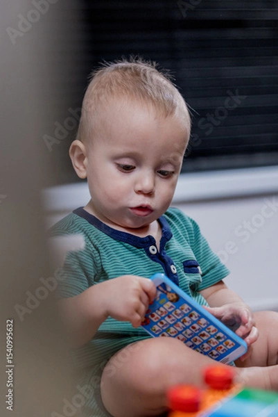 Fototapeta A small baby is happily sitting on the floor and playing with a toy phone