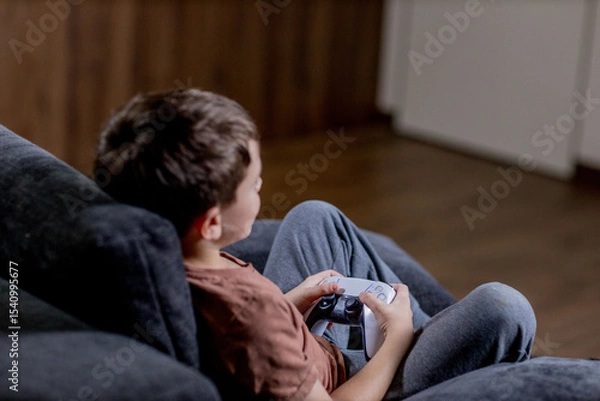 Fototapeta Boy is sitting on sofa at home and playing video games with his joypad.
