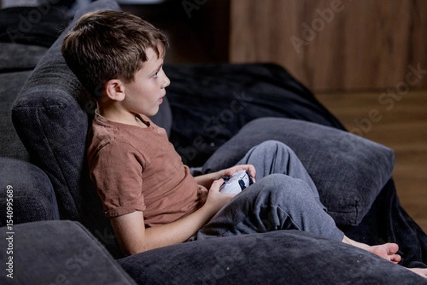 Fototapeta Boy is sitting on sofa at home and playing video games with his joypad.