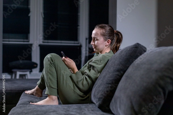 Fototapeta A woman is comfortably sitting on a cozy couch while looking in her phone