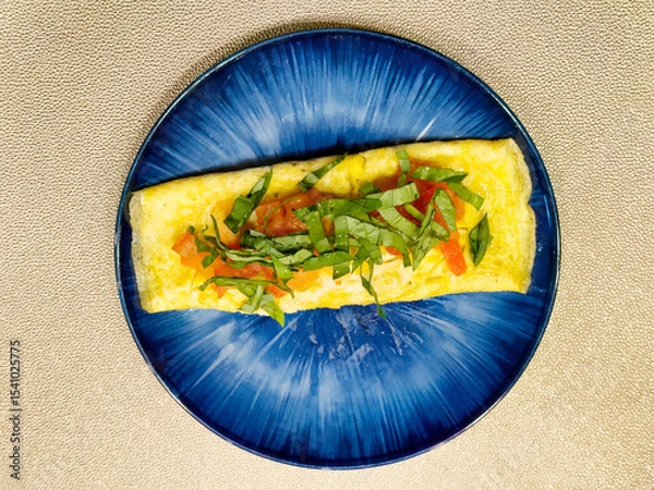 Fototapeta Morning breakfast meal of a large egg omelet with sliced tomato and basil on dark blue plate in overhead flat lay view