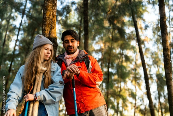 Fototapeta Young Couple Resting Against Tree During a Winter Hike. Enjoying the Golden Hour Light in a Peaceful Forest, Wearing Warm Clothes and Backpacks, Sharing a Quiet and Happy Moment Together
