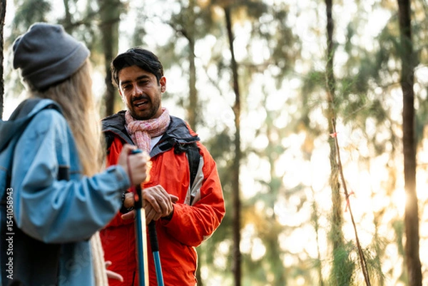Fototapeta Young Couple Resting Against Tree During a Winter Hike. Enjoying the Golden Hour Light in a Peaceful Forest, Wearing Warm Clothes and Backpacks, Sharing a Quiet and Happy Moment Together