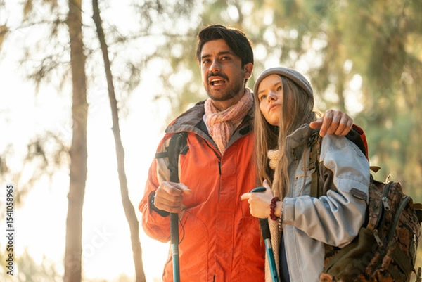 Fototapeta Romantic Couple Hiking in the Forest During Golden Hour. Embracing While Watching Sunset Light Through Trees, Wearing Warm Jackets and Backpacks, Feeling Connected and Adventurous in Nature
