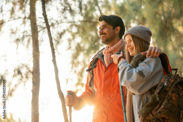 Fototapeta Romantic Couple Hiking in the Forest During Golden Hour. Embracing While Watching Sunset Light Through Trees, Wearing Warm Jackets and Backpacks, Feeling Connected and Adventurous in Nature