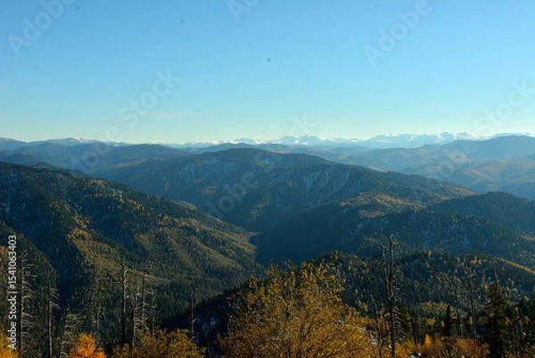 Obraz View from the top of the mountain ranges covered with dense coniferous forest and snow-capped mountains in the distance on a sunny autumn evening.