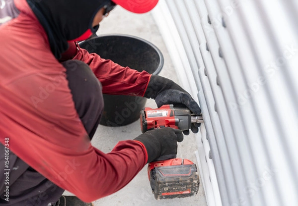 Fototapeta The worker is using a cordless impact wrench to tighten bolts on the metal sheet tank wall .