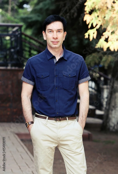 Obraz Молодой красивый брюнет мужчина позирует в синей рубашке, светлых летних штанах и часах
