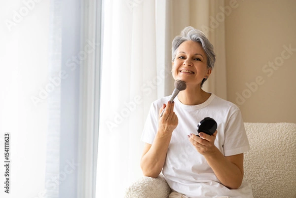 Fototapeta Smiling senior woman applying makeup with brush while sitting in armchair in bright home interior