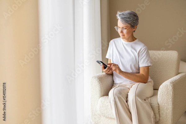 Fototapeta Senior woman using smartphone at home sitting in soft armchair near window in modern bright room