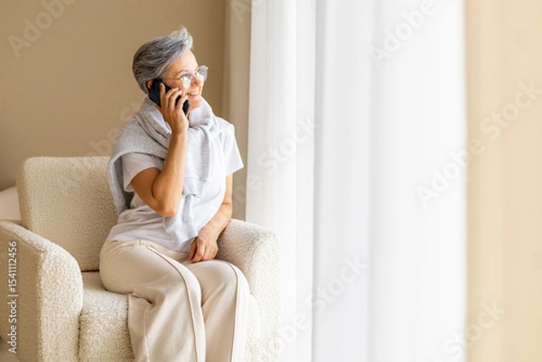 Fototapeta Smiling mature woman talking on smartphone while sitting on cozy armchair by bright window light