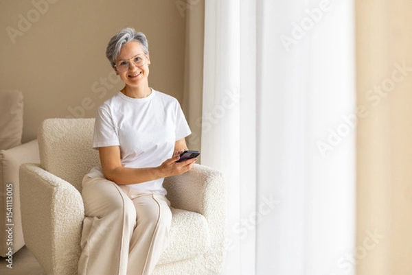 Fototapeta Smiling senior woman using smartphone at home in cozy modern interior with soft natural light