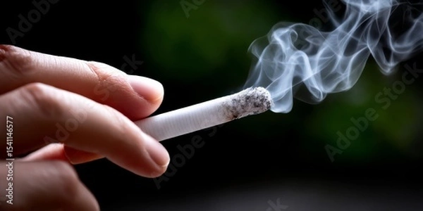 Fototapeta Smoking Cigarette: A close-up shot of a hand holding a burning cigarette, smoke curling upwards against a dark, blurred background.