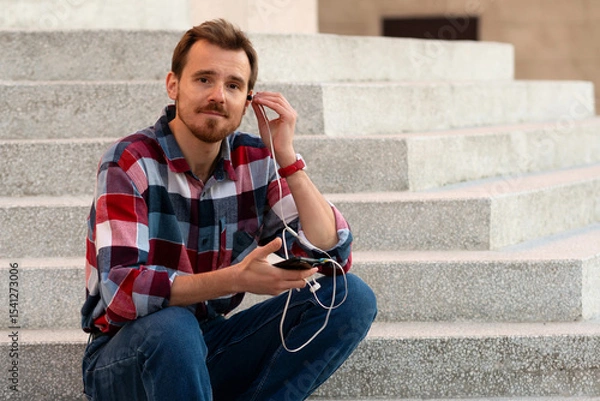 Fototapeta Man seated on steps adjusting earphones connected to smartphone. Guy with beard and modern hairstyle wearing in blue, red and white plaid shirt walking through the city streets in urban background