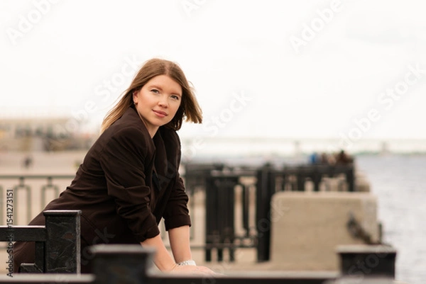 Fototapeta Smiling woman in brown blazer looking at camera near waterfront. Natural feel with soft light. Casual, confident pose and expression add genuine and approachable vibe