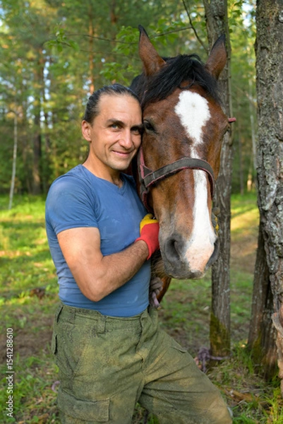 Obraz rustic man with  horse in rural village near  forest.  horse farm is surrounded by nature, hay is stacked nearby,  peaceful and traditional.  man is caring for  horse