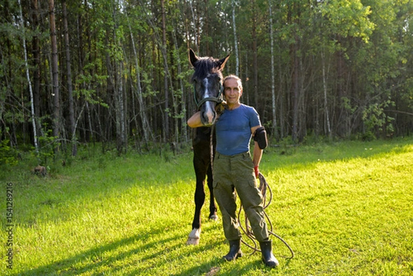 Obraz rustic man with  horse in rural village near  forest.  horse farm is surrounded by nature, hay is stacked nearby,  peaceful and traditional.  man is caring for  horse