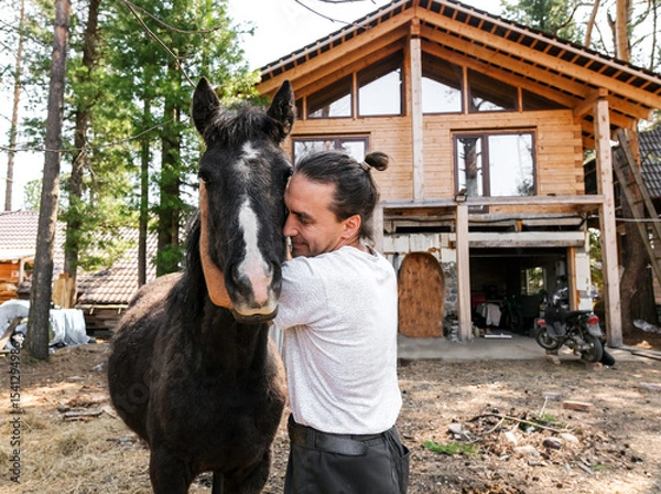 Obraz rustic man with  horse in rural village near  forest.  horse farm is surrounded by nature, hay is stacked nearby,  peaceful and traditional.  man is caring for  horse