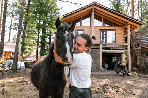 Obraz rustic man with  horse in rural village near  forest.  horse farm is surrounded by nature, hay is stacked nearby,  peaceful and traditional.  man is caring for  horse