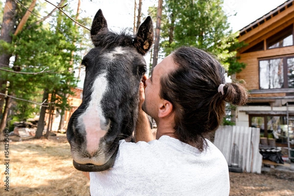Obraz rustic man with  horse in rural village near  forest.  horse farm is surrounded by nature, hay is stacked nearby,  peaceful and traditional.  man is caring for  horse