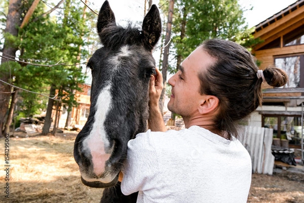 Obraz rustic man with  horse in rural village near  forest.  horse farm is surrounded by nature, hay is stacked nearby,  peaceful and traditional.  man is caring for  horse