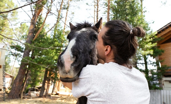Obraz rustic man with  horse in rural village near  forest.  horse farm is surrounded by nature, hay is stacked nearby,  peaceful and traditional.  man is caring for  horse