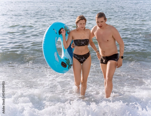 Fototapeta The guy carefully leads the girl out of the water onto the shore after she was covered by a wave. She walks with her eyes closed from the sea water trapped in them.