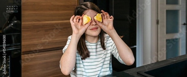Fototapeta Banner of young girl having fun in the kitchen, holding heart shaped cookies over her eyes, expressing playfulness and enjoyment. Happy little girl looking at camera while covering eyes with biscuits.