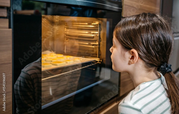 Fototapeta Little girl watching cookies baking in oven, waiting for them to be ready, enjoying the sweet smell and anticipating the delicious taste. Cooking at leisure time concept. Left copy space available.