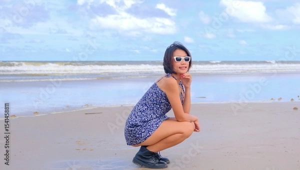 Obraz Pretty Asian woman in a floral sundress and black ankle boots, crouching on a beach against a serene ocean backdrop