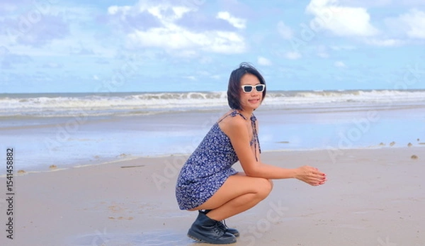 Obraz Pretty Asian woman in a floral sundress and black ankle boots, crouching on a beach against a serene ocean backdrop