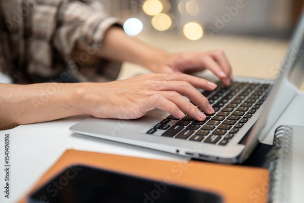 Fototapeta Close up of a man's both hands typing on laptop with phone atop book sitting at white table in cafe.