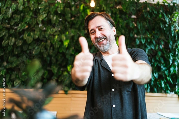 Fototapeta Happy middle-aged cafe owner giving two thumbs up in front of a lush green wall. He smiles confidently, celebrating business success and customer satisfaction in a cozy, eco-friendly setting.