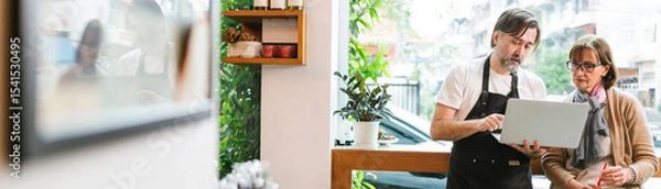 Fototapeta Cafe owner and financial advisor discussing business strategy using a laptop in a modern coffee shop. Bright setting with plants and wine bottles, symbolizing collaboration and entrepreneurship.