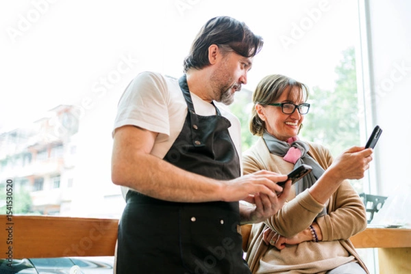 Fototapeta Cafe owner and business advisor discussing marketing or finance using smart phones. Both are smiling and engaged, symbolizing teamwork, strategy, and modern small business collaboration.