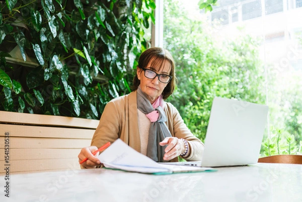 Fototapeta Mature woman working at a cafe with a laptop and financial documents. She reviews papers intently in a green, cozy setting, symbolizing focus, professionalism, and modern entrepreneurship.