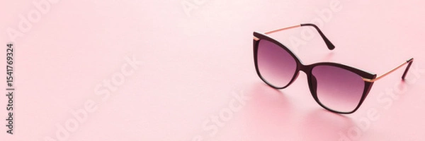 Fototapeta Female sunglasses with dark black plastic frame on light pink table background. Pastel color. Closeup. Wide banner. Empty place for text.