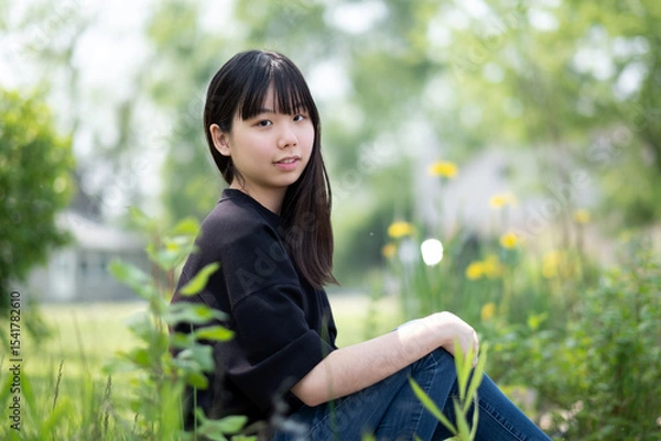 Fototapeta Young asian teenage girl sitting on the ground in a grassy area, surrounded by green plants and wildflowers, wearing a black top and jeans.
