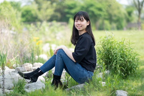 Fototapeta Young asian teenage girl sitting on the ground in a grassy area, surrounded by green plants and wildflowers, wearing a black top and jeans.