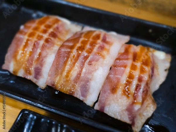 Fototapeta Grilled bacon-wrapped sushi served on a black tray. Close-up image highlighting the juicy texture, glossy sauce, and savory Japanese fusion style presentation.