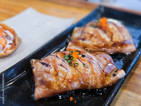 Fototapeta Grilled salmon sushi belly topped with fish roe and chopped scallions, glazed with teriyaki sauce and served on a black ceramic plate