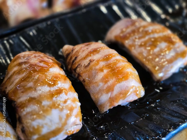 Fototapeta Grilled salmon nigiri made from belly cut with skin-on, glazed with teriyaki sauce and served on a black plate. Rich texture and glossy finish highlight freshness.
