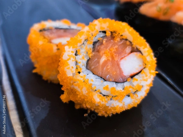 Fototapeta Sushi rolls topped with vibrant orange tobiko, filled with salmon and crab stick, served on a black ceramic plate in Japanese restaurant setting