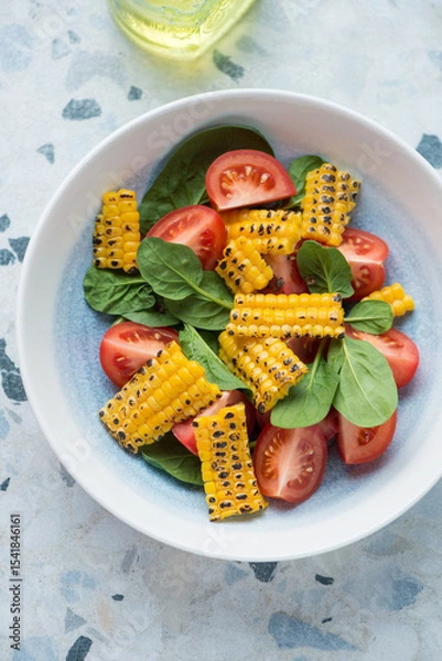 Fototapeta Plate with tomato, fresh spinach and grilled corn salad, vertical shot on a white and blue granite background, high angle view