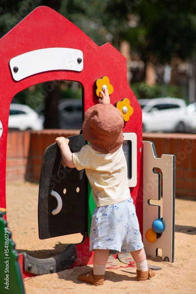 Obraz A toddler wearing a sun hat and summer clothes is engaged at an outdoor playground. The child is interacting with a colourful play structure featuring a red panel. Lifestyle photo, summer time.