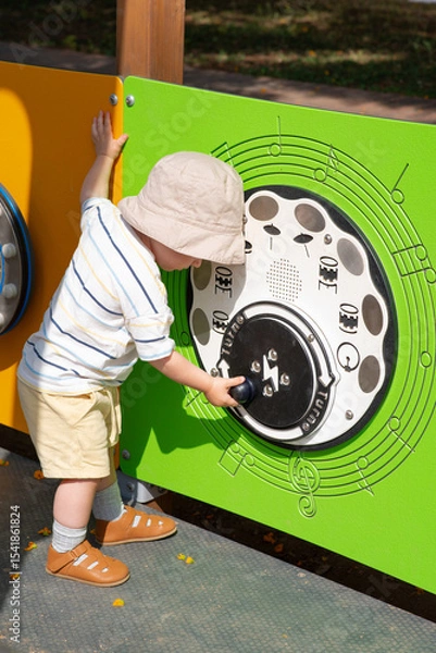 Obraz Toddler in sun hat and summer clothes playing with an interactive musical panel with sounds of drums at playground. The child is turning a dial, engaged in sensory and motor development activity. 