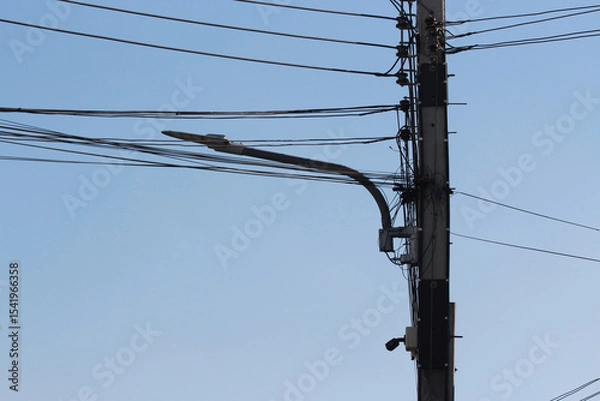 Fototapeta Streetlight and Wires: A close-up view of a streetlight mounted on a utility pole, surrounded by a complex network of electrical wires against a clear blue sky.