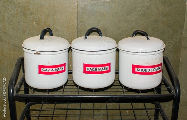 Obraz iron rack placed outside an ICU in a hospital, containing three white tin boxes labeled Cap and Mask, Face Mask, and Shoe Cover. reflects hospital hygiene protocols and infection control measures.